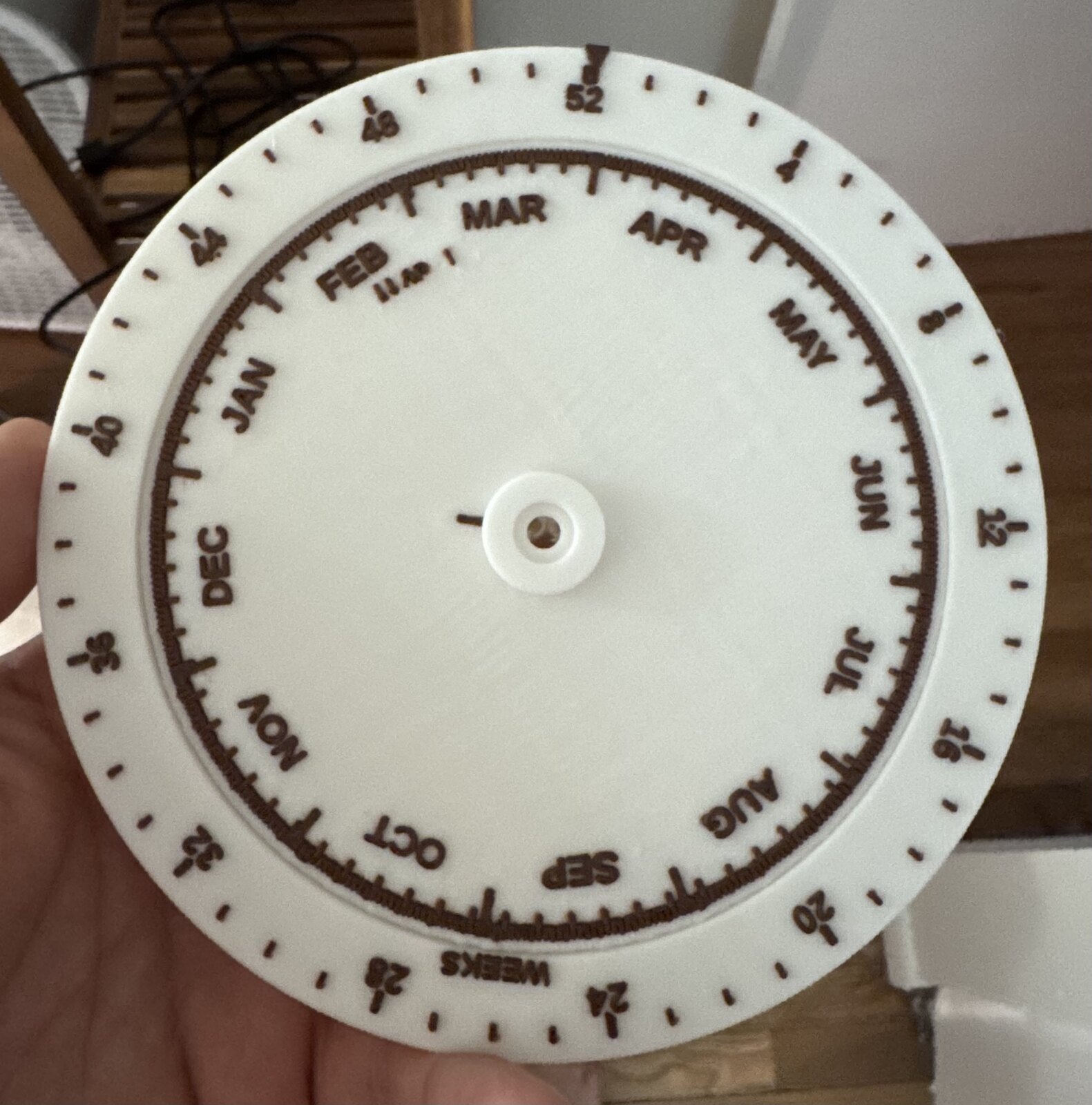 Photo of the first print held in one hand: a white disc with espresso-brown embossed markings. The inner ring carries JAN through DEC, the outer rim is numbered in weeks from 4 to 52, and a small index arrow sits at the top